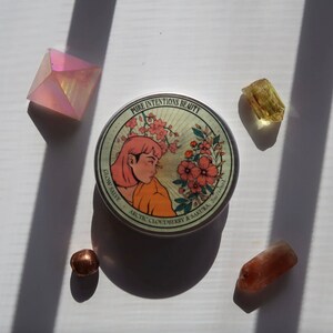 May include: A round metal container with the text "Pure Intentions Beauty" and a graphic of a woman with pink hair and flowers. The container is surrounded by crystals and a copper-colored bead. The product is 2oz/60ml.