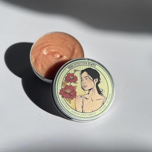 May include: Open metal tin with a peach-colored solid serum. The lid features a woman's illustration with flowers and the text "PURE INTENTIONS BEAUTY, AMATERASU AGELESS SOLID SERUM, TSUBAKI 1oz/30ml."