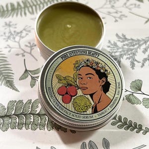 May include: A small, open metal tin containing a green solid serum, with a second tin in the foreground. The front tin has a label with the text "PURE INTENTIONS BEAUTY" and "RESTING GODDESS FACE SOLID SERUM". The label features a woman's portrait.