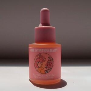 Puede incluir: Botella de vidrio rosa de Glow Oil Serum con cuentagotas. La etiqueta presenta el texto "Pure Intentions Beauty" y una ilustraci&oacute;n de una mujer y flores. La botella contiene 20 ml de producto.