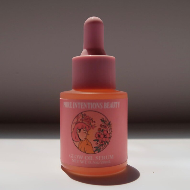 Puede incluir: Botella de vidrio rosa de Glow Oil Serum con cuentagotas. La etiqueta presenta el texto "Pure Intentions Beauty" y una ilustraci&oacute;n de una mujer y flores. La botella contiene 20 ml de producto.