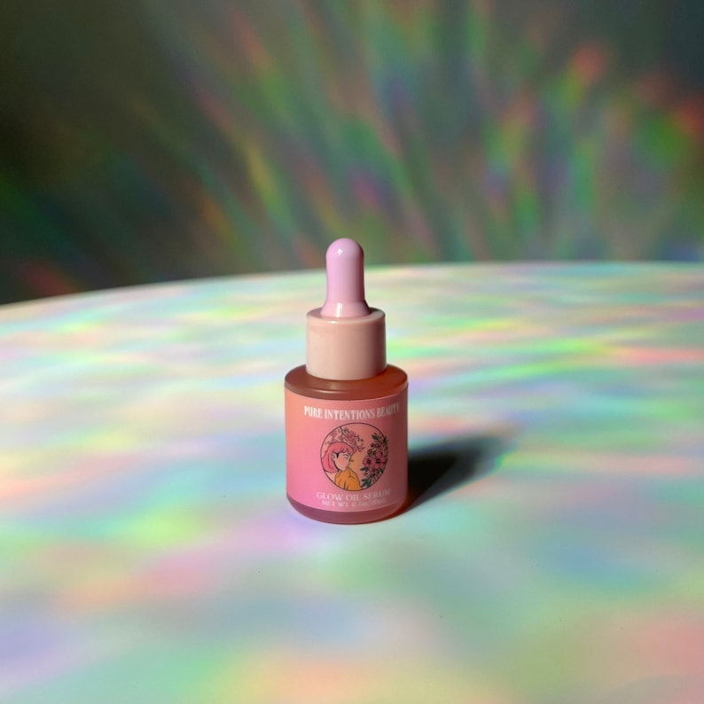 Puede incluir: Una peque&ntilde;a botella de vidrio rosa de Glow Oil Serum con cuentagotas. La botella tiene una tapa rosa y una etiqueta con el texto "Pure Intentions Beauty" y una ilustraci&oacute;n floral. El fondo es una superficie brillante e iridiscente.