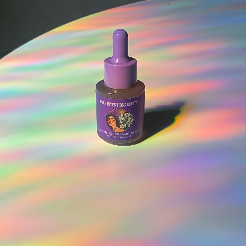 May include: A small, clear glass bottle with a purple dropper and cap. The label reads "Pure Intentions Beauty" and "Mangosteen Dreams Oil Serum". The bottle is set against a background with rainbow light reflections.