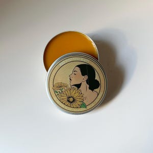 Pu&ograve; includere: Un barattolo rotondo di metallo aperto con un balsamo dorato all'interno. Il coperchio presenta il profilo di una donna, girasoli e il testo "PURE INTENTIONS BEAUTY AU REVOIR ACNE CLEANSING BALM". Il balsamo &egrave; di un giallo vivace.