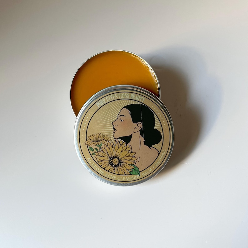 Pu&ograve; includere: Un barattolo rotondo di metallo aperto con un balsamo dorato all'interno. Il coperchio presenta il profilo di una donna, girasoli e il testo "PURE INTENTIONS BEAUTY AU REVOIR ACNE CLEANSING BALM". Il balsamo &egrave; di un giallo vivace.