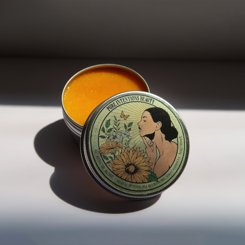 May include: Open metal tin with a bright orange balm. The lid features a woman's profile, a butterfly, and sunflowers, with the text "PURE INTENTIONS BEAUTY". The balm is labeled "AU REVOIR ACNE - MANGO BUTTER, SEA BUCKTHORN, AND CALENDULA 1oz/30ml".