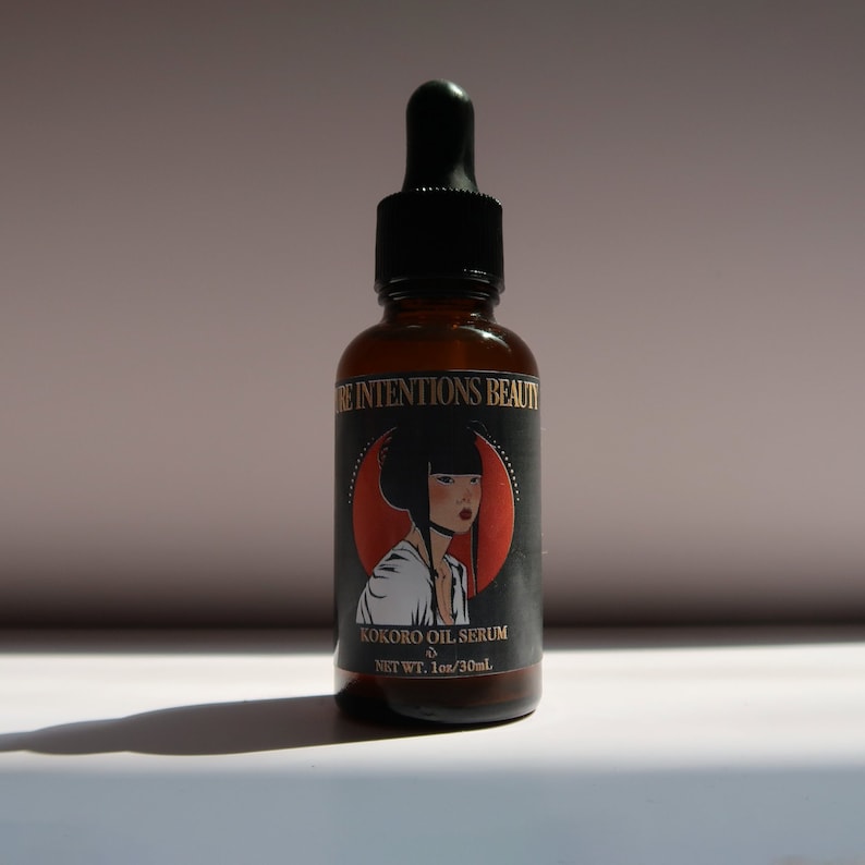 May include: A brown glass bottle with a black dropper and a label featuring a woman's portrait. The label reads "PURE INTENTIONS BEAUTY" and "KOKORO OIL SERUM" with a net weight of 1 fl oz / 30ml.