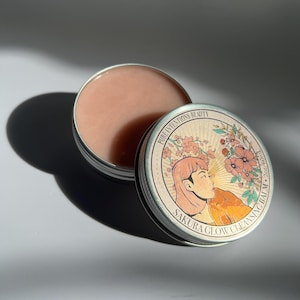 May include: An open, round, silver-colored tin of Sakura Glow Cleansing Balm. The balm is a light pink color. The lid, next to the tin, has a vintage-style illustration of a woman and flowers, with the text "Sakura Glow Cleansing Balm".
