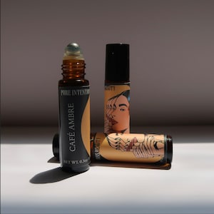 May include: Three amber glass roll-on bottles. One bottle is upright, labeled "CAFÉ AMBRE" and "PURE INTENTION". The other two bottles have a graphic of a woman's face. The bottles are on a white surface.