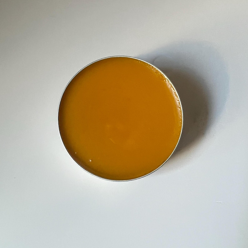Pu&ograve; includere: Un barattolo rotondo d'argento riempito con un balsamo liscio giallo dorato. Il balsamo ha un aspetto lucido ed &egrave; centrato su una superficie bianca. La presentazione generale &egrave; pulita e semplice, evidenziando il colore e la consistenza del prodotto.