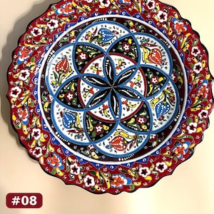 May include: A round ceramic plate with a scalloped edge, featuring a central geometric pattern of intersecting black and white arcs. The design includes colorful floral motifs in red, blue, yellow, and green. The border is a vibrant red.