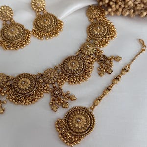 May include: A gold-toned jewelry set featuring a necklace, earrings, and a headpiece. The set has a floral design with intricate detailing and small dangling elements. The jewelry is displayed on a white surface.