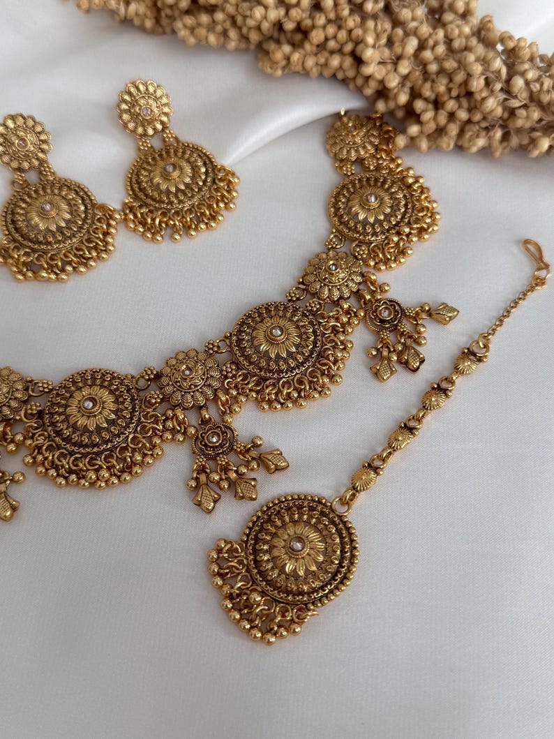 May include: A gold-toned jewelry set featuring a necklace, earrings, and a headpiece. The set has a floral design with intricate detailing and small dangling elements. The jewelry is displayed on a white surface.