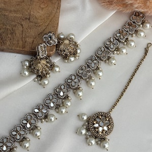 Puede incluir: Un conjunto de joyas de color oro antiguo con collar, pendientes y diadema. El conjunto incluye dise&ntilde;os florales con detalles de espejo y perlas de imitaci&oacute;n colgantes. Los pendientes tienen forma de campana. Las joyas se exhiben sobre tela blanca.