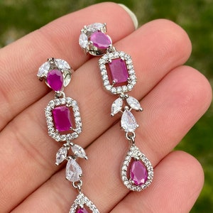 May include: A pair of silver dangle earrings with pink gemstones and clear cubic zirconia stones. The earrings feature a teardrop-shaped stone at the bottom, a square stone in the middle, and a smaller teardrop-shaped stone at the top.