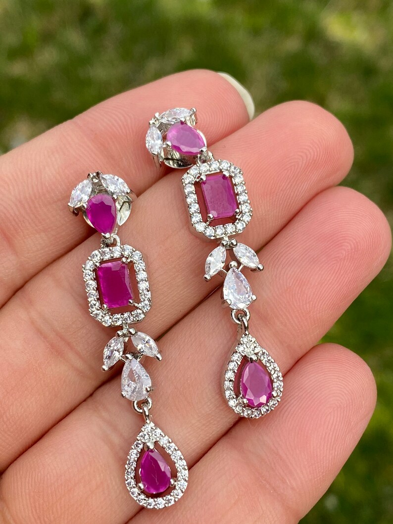 May include: A pair of silver dangle earrings with pink gemstones and clear cubic zirconia stones. The earrings feature a teardrop-shaped stone at the bottom, a square stone in the middle, and a smaller teardrop-shaped stone at the top.