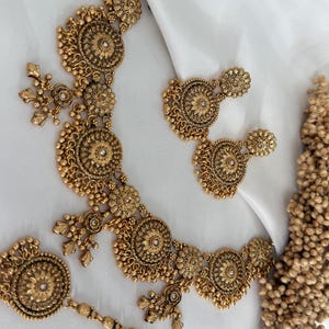 May include: A gold-toned jewelry set featuring a necklace, earrings, and a headpiece. The set has intricate floral and circular designs with small, pearl-like accents. The pieces are arranged on a white surface.