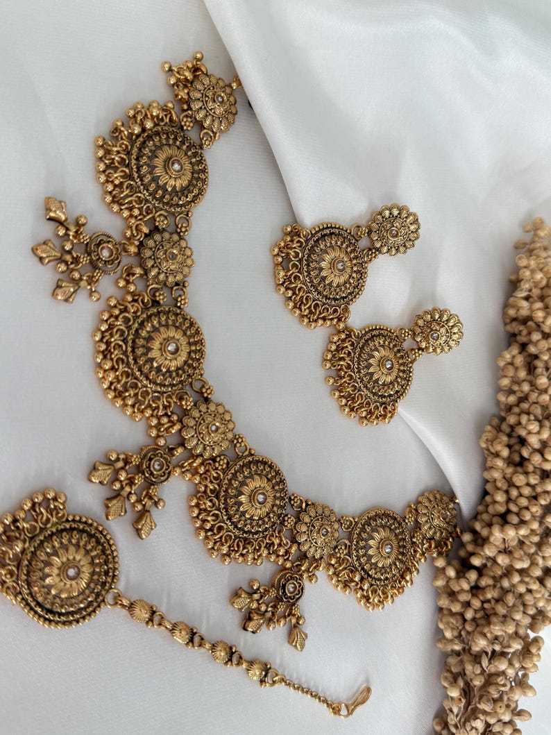 May include: A gold-toned jewelry set featuring a necklace, earrings, and a headpiece. The set has intricate floral and circular designs with small, pearl-like accents. The pieces are arranged on a white surface.