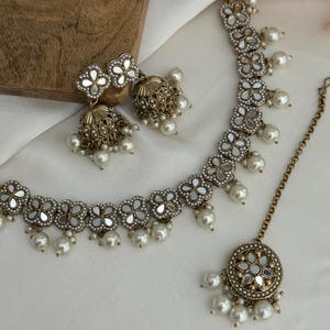 Puede incluir: Un conjunto de joyas de tres piezas que incluye un collar, pendientes y una pieza para la cabeza. El conjunto tiene un acabado plateado con detalles florales de espejo y gotas de perlas de imitaci&oacute;n. Los pendientes tienen forma de campana. El collar y la pieza para la cabeza tienen un dise&ntilde;o floral a juego.