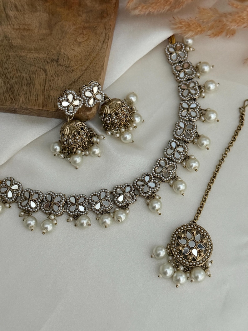 Puede incluir: Un conjunto de joyas de tres piezas que incluye un collar, pendientes y una pieza para la cabeza. El conjunto tiene un acabado plateado con detalles florales de espejo y gotas de perlas de imitaci&oacute;n. Los pendientes tienen forma de campana. El collar y la pieza para la cabeza tienen un dise&ntilde;o floral a juego.