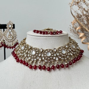 May include: A gold and white necklace with red beads. The necklace has a large, ornate design with multiple round, circular elements. The necklace is on a white mannequin.