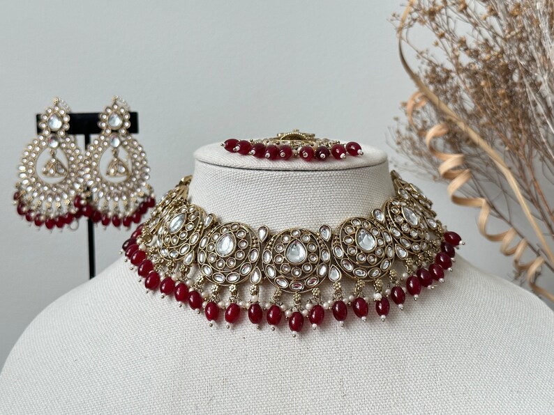 May include: A gold and white necklace with red beads. The necklace has a large, ornate design with multiple round, circular elements. The necklace is on a white mannequin.
