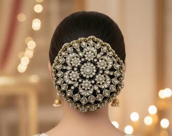 May include: A circular hair bun cover featuring an intricate gold-tone metal filigree design adorned with numerous clear white crystals and stones. The accessory is centered over a dark hair bun, with small gold-tone bell-shaped earrings visible.