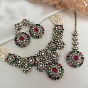 May include: An elaborate jewellery set comprising a necklace, earrings, and a headpiece. The silver-toned pieces are embellished with colourful gemstones in red, green, and blue, and surrounded by small white stones. The necklace has a pearl strand.