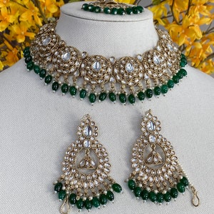 May include: A gold necklace and earrings set with a green and white beaded design. The necklace features a large, ornate pendant with a circular design and the earrings are teardrop shaped.