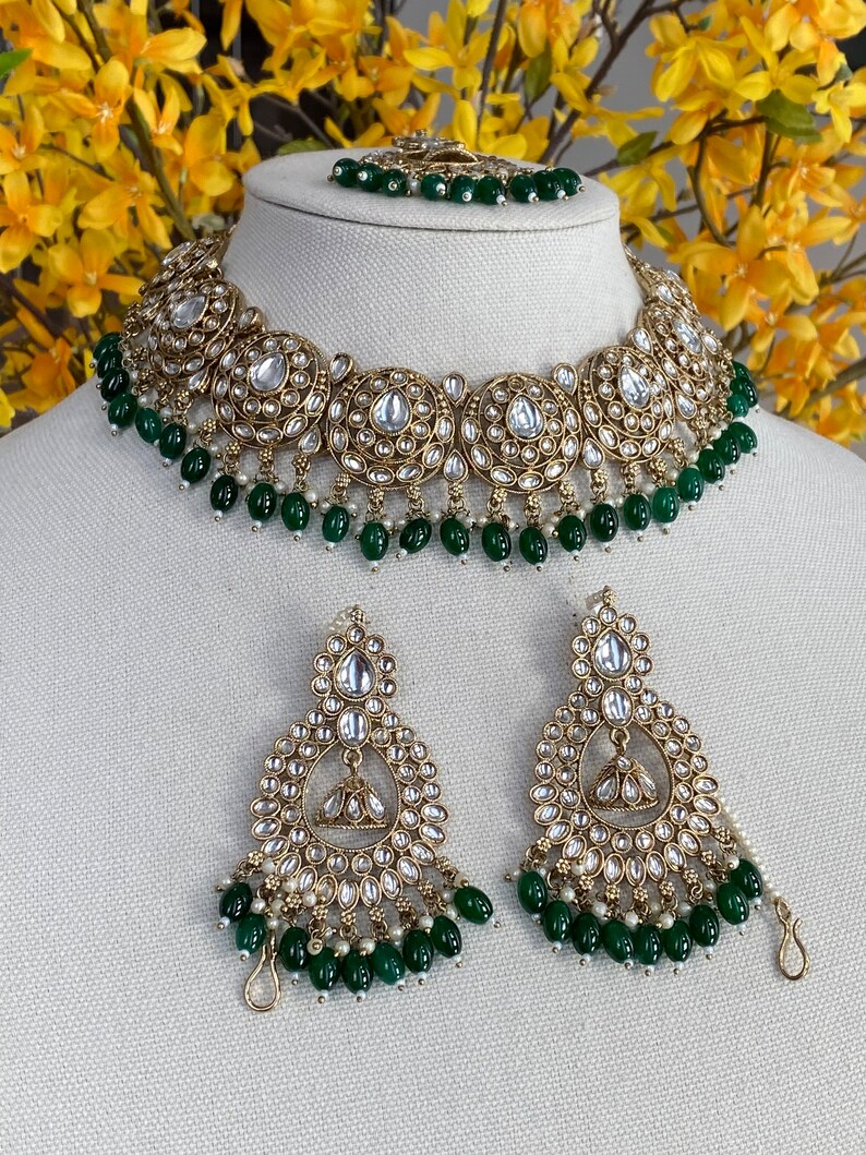 May include: A gold necklace and earrings set with a green and white beaded design. The necklace features a large, ornate pendant with a circular design and the earrings are teardrop shaped.