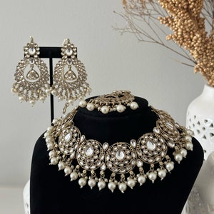 May include: A gold and white bridal necklace and earrings set with pearl accents. The necklace features a large, round design with multiple smaller circles and clear stones. The earrings are teardrop shaped with similar embellishments.