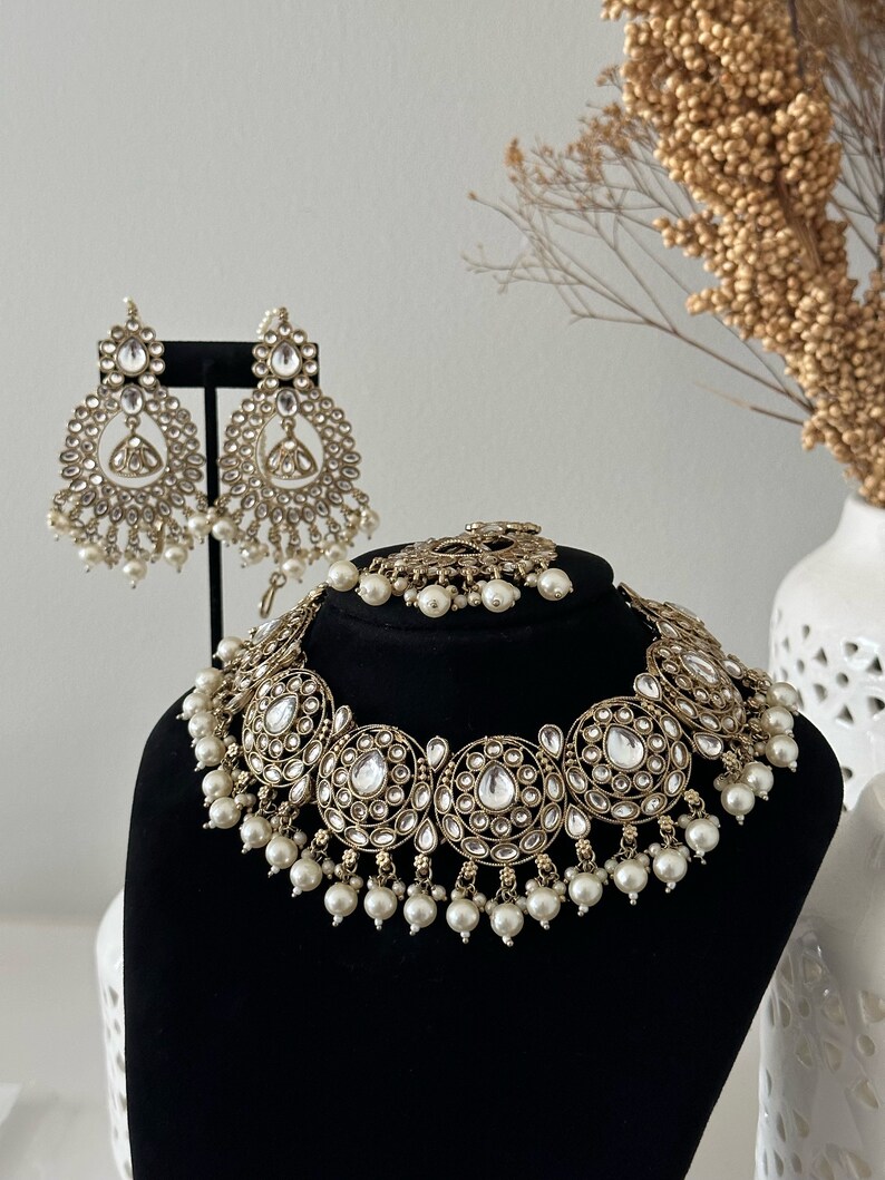 May include: A gold and white bridal necklace and earrings set with pearl accents. The necklace features a large, round design with multiple smaller circles and clear stones. The earrings are teardrop shaped with similar embellishments.