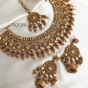 以下が含まれることがあります： アンティークゴールドトーンのジュエリーセット。ネックレス、イヤリング、ヘッドピースが含まれています。セットは、小さな透明な宝石が付いた、複雑な花柄とビーズのデザインです。ジュエリーは、白くテクスチャ加工された表面に表示されています。「TAZORI COLLECTION」のブランド名が見えます。