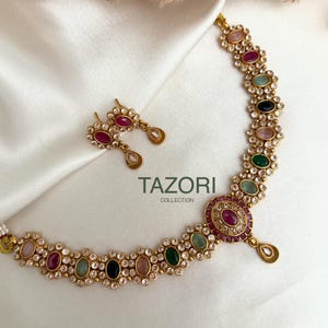 Peut inclure: Un collier doré et une paire de boucles d'oreilles assorties. Le collier et les boucles d'oreilles présentent un motif floral avec une variété de pierres précieuses colorées, notamment des pierres roses, rouges, vertes et noires, entourées de cristaux transparents. Le texte "TAZORI COLLECTION" est visible.