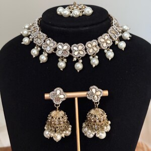 Puede incluir: Un conjunto de joyer&iacute;a de tres piezas que incluye un collar gargantilla, un par de pendientes y un accesorio para el cabello. El conjunto incluye un collar plateado con detalles de espejo en forma de flor y perlas de imitaci&oacute;n colgantes. Los pendientes son dorados, en forma de campana y adornados con perlas.