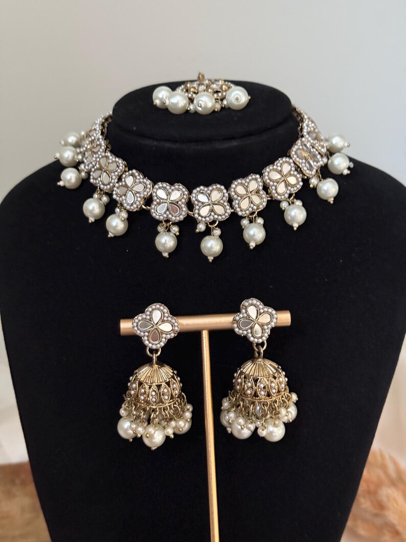 Puede incluir: Un conjunto de joyer&iacute;a de tres piezas que incluye un collar gargantilla, un par de pendientes y un accesorio para el cabello. El conjunto incluye un collar plateado con detalles de espejo en forma de flor y perlas de imitaci&oacute;n colgantes. Los pendientes son dorados, en forma de campana y adornados con perlas.