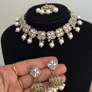 Puede incluir: Un conjunto de joyas plateadas que incluye un collar, pendientes y una pieza para la cabeza. El collar y los pendientes tienen un dise&ntilde;o floral con centros espejados y perlas blancas colgantes. Los pendientes tienen forma de campana con detalles de perlas.