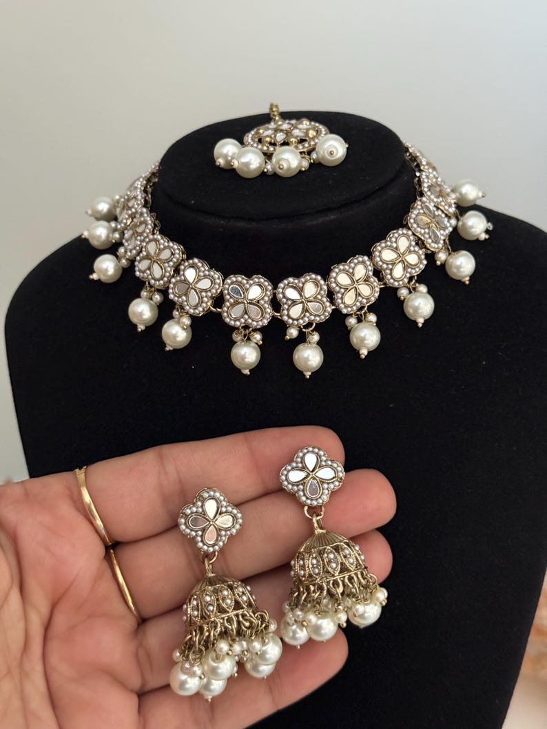 Puede incluir: Un conjunto de joyas plateadas que incluye un collar, pendientes y una pieza para la cabeza. El collar y los pendientes tienen un dise&ntilde;o floral con centros espejados y perlas blancas colgantes. Los pendientes tienen forma de campana con detalles de perlas.
