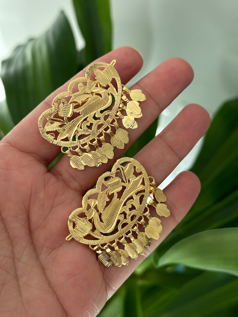 May include: A pair of gold filigree hair clips with a heart-shaped design and intricate floral patterns. The clips have a dangling fringe of small, round shapes.