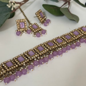 May include: A purple and gold choker necklace with a dangling design of purple beads and white pearls. The necklace comes with a matching pair of earrings with purple stones and white pearls.