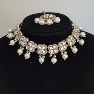Puede incluir: Un collar plateado y un conjunto de tocado a juego. El collar presenta colgantes en forma de flor con centros espejados y perlas de imitaci&oacute;n colgantes. El tocado tiene un dise&ntilde;o similar con un grupo de perlas.