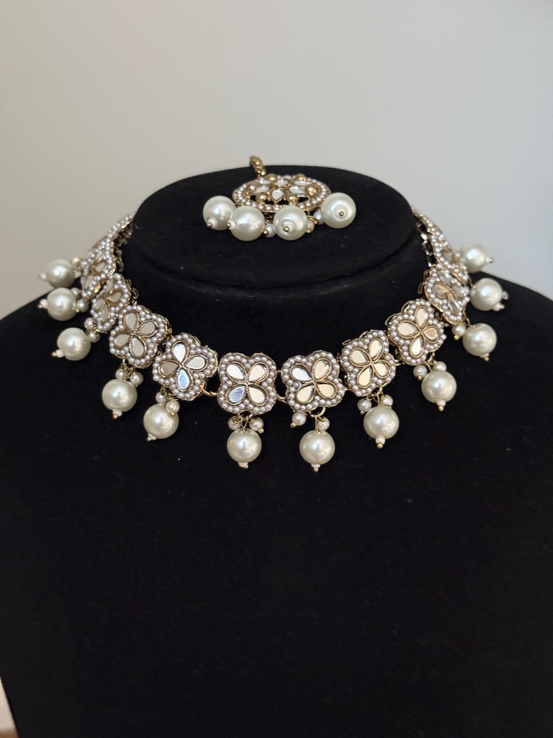 Puede incluir: Un collar plateado y un conjunto de tocado a juego. El collar presenta colgantes en forma de flor con centros espejados y perlas de imitaci&oacute;n colgantes. El tocado tiene un dise&ntilde;o similar con un grupo de perlas.