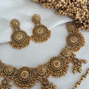 May include: A gold-toned jewelry set featuring a necklace and matching earrings. The necklace and earrings have a floral design with small dangling beads. The jewelry is displayed on a white surface, with dried flowers in the background.