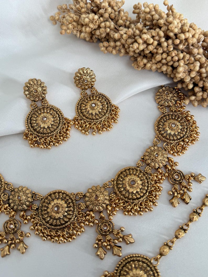 May include: A gold-toned jewelry set featuring a necklace and matching earrings. The necklace and earrings have a floral design with small dangling beads. The jewelry is displayed on a white surface, with dried flowers in the background.
