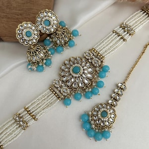 Peut inclure: Un ensemble de collier et boucles d'oreilles en or et blanc avec des perles bleues. Le collier est de style ras de cou avec plusieurs rangs de perles blanches et des accents dorés. Les boucles d'oreilles sont de style pendantes avec un grand pendentif rond et des perles bleues. Le maang tikka est une coiffure indienne traditionnelle avec un grand pendentif rond et des perles bleues.
