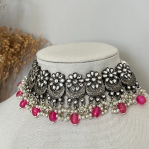 May include: A silver choker necklace with a floral design and pink beads. The necklace has a silver chain with a floral design and is adorned with small white stones. The necklace is finished with a row of pink beads.