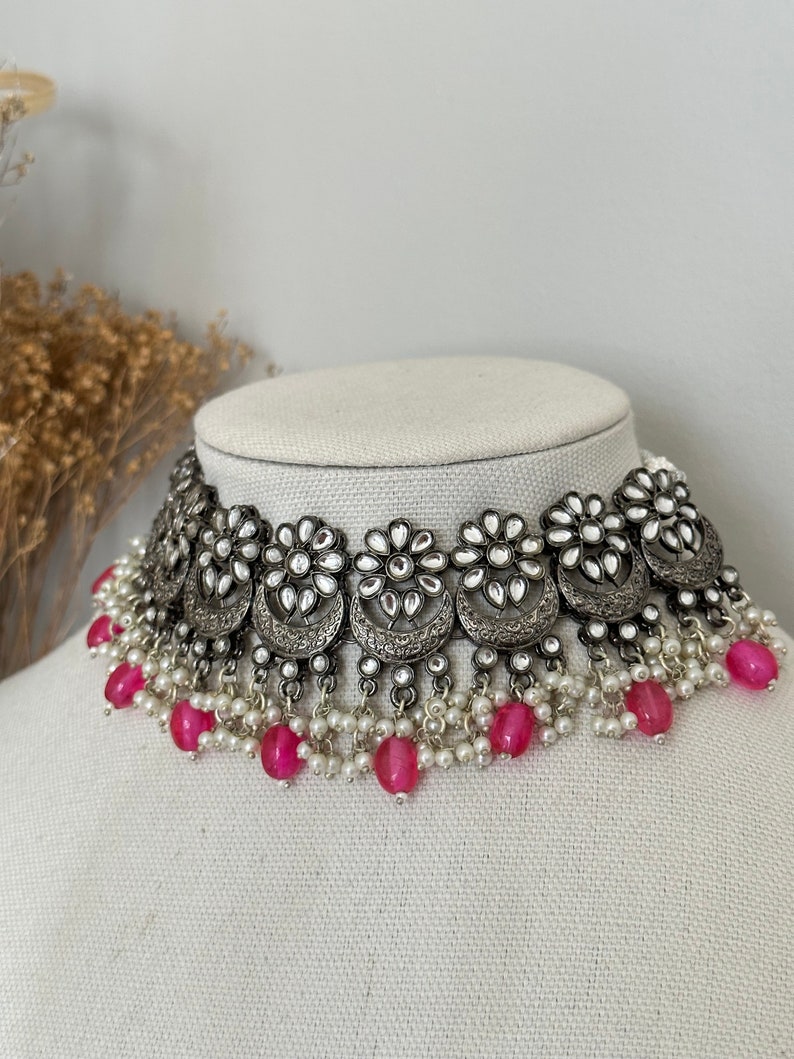 May include: A silver choker necklace with a floral design and pink beads. The necklace has a silver chain with a floral design and is adorned with small white stones. The necklace is finished with a row of pink beads.