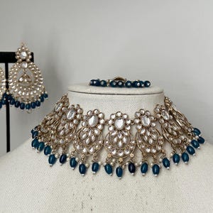 May include: A gold and white necklace with blue beads. The necklace is on a white mannequin. The necklace has a large, ornate pendant in the center.