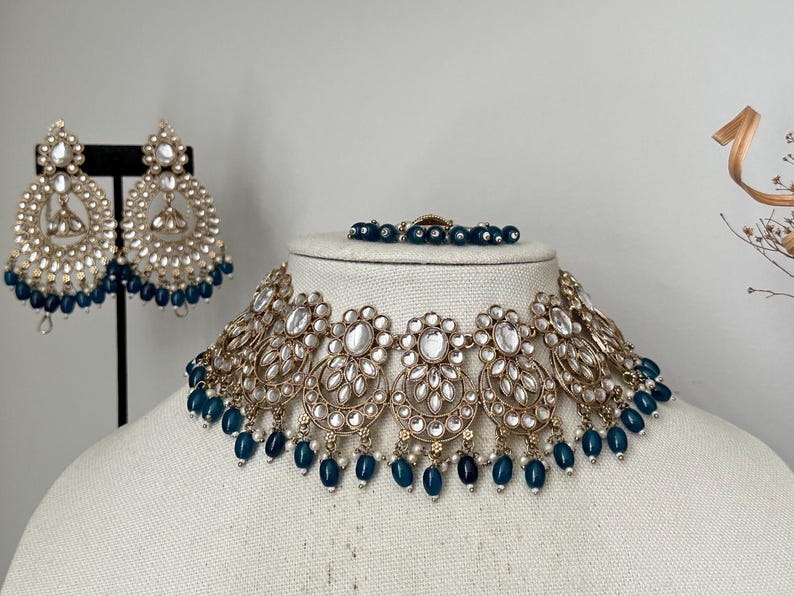 May include: A gold and white necklace with blue beads. The necklace is on a white mannequin. The necklace has a large, ornate pendant in the center.