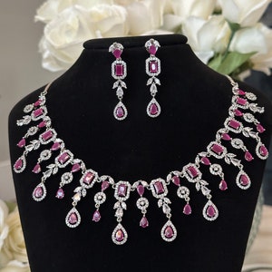 May include: A silver necklace and earrings set with pink cubic zirconia stones. The necklace features a delicate chain with a pendant design and the earrings are drop style.